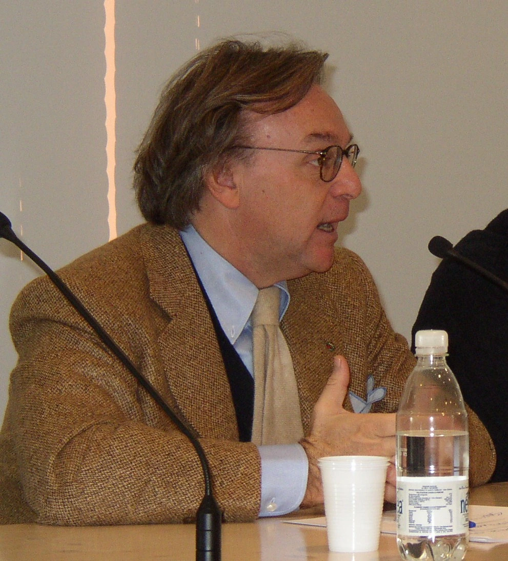 Diego Della Valle speaking at a meeting, in profile, wearing glasses and a jacket.