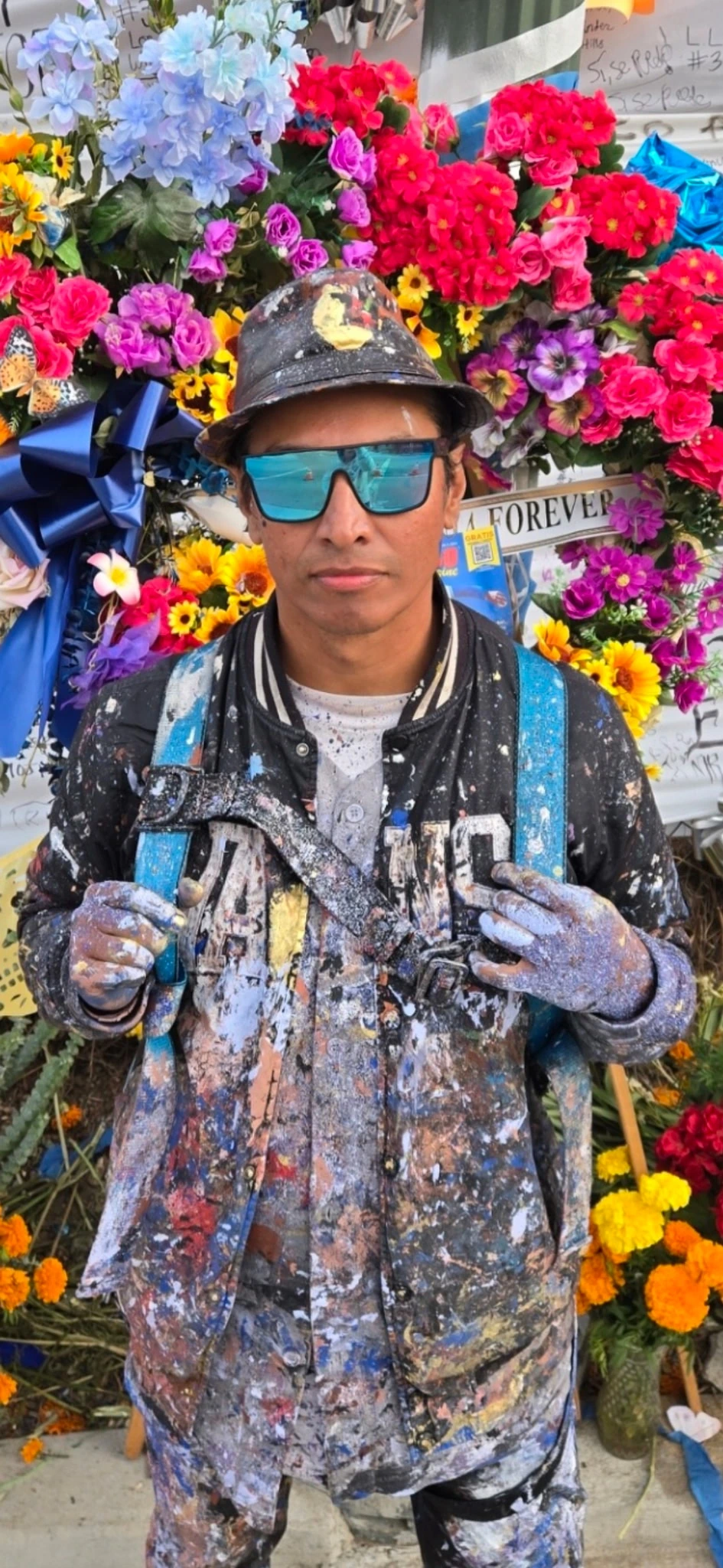 Robert Vargas standing in paint-splattered work clothes in front of a colorful floral mural backdrop
